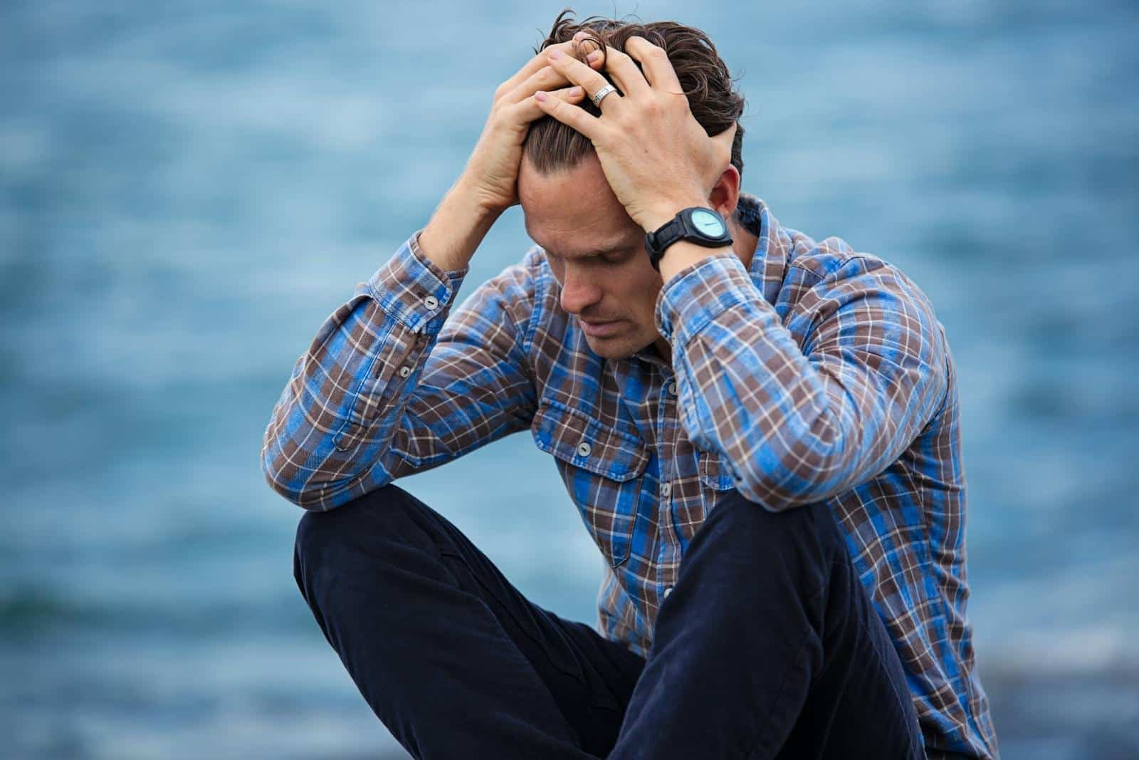 Man in plaid shirt lost in thought by calm water, reflecting inner turmoil and introspection.