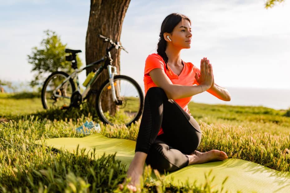 Slim beautiful woman doing sports in morning in park doing yoga on mat in colorful fitness outfit in nature, smiling happy healthy lifestyle, calm meditation, bicycle on background