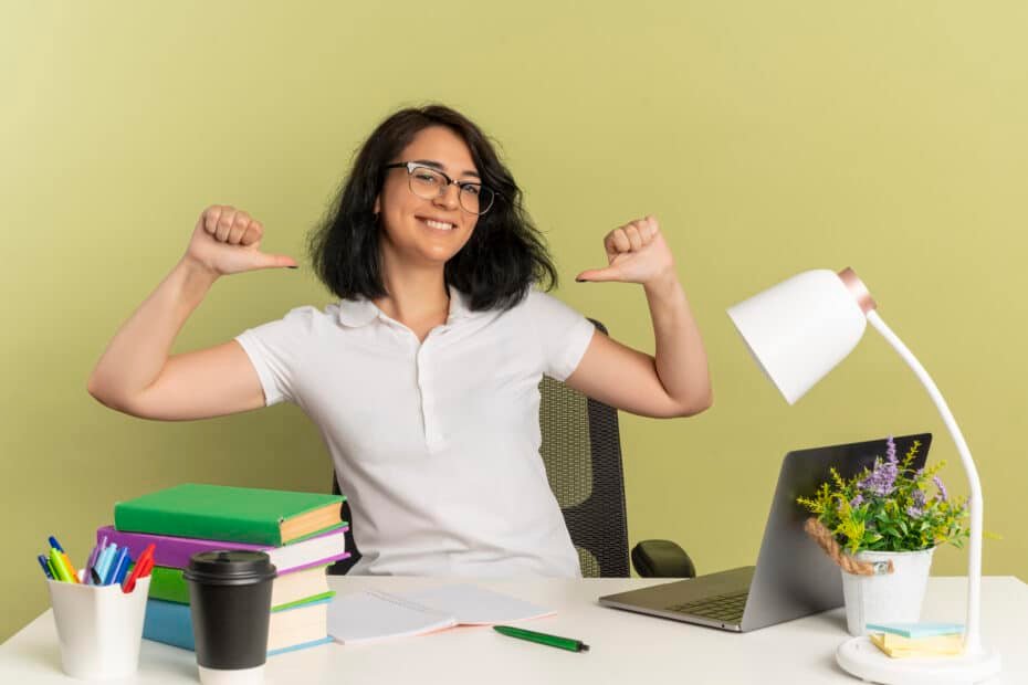 Young smiling pretty caucasian schoolgirl wearing glasses sits at desk with school tools points at herself isolated on green background with copy space